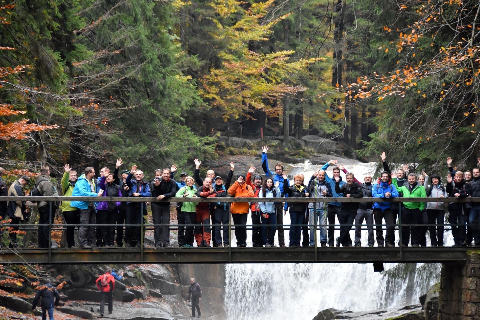 Skupina v outdoorovém oblečení mává na lávce nad vodopádem v horách